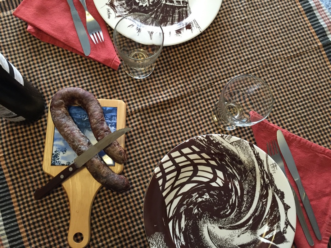 Fotografia d'una taula parada per dos: hi ha dos plats, dues copes, una fusta amb una botifarra al damunt i una ampolla de vi negre. Les estovalles són de farcell